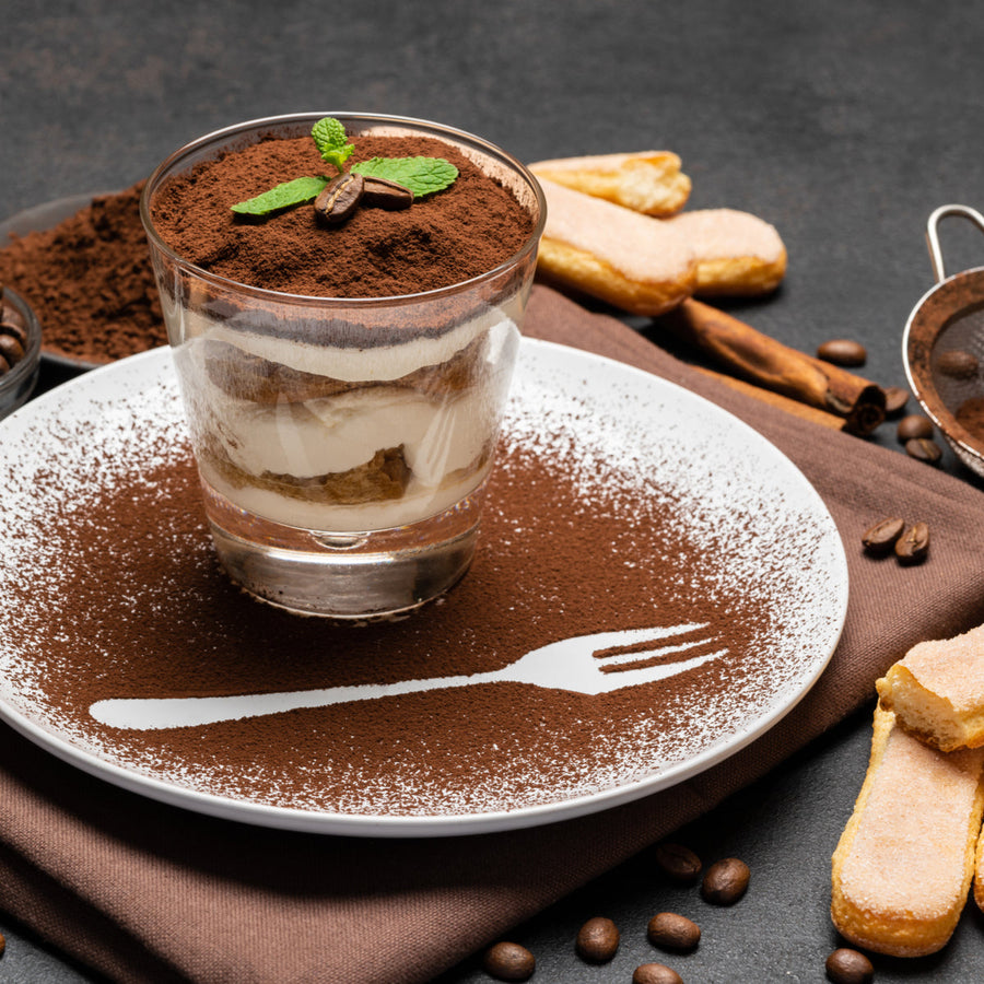 A glass dessert filled with layers of cream and cake is topped with cocoa powder and a mint leaf, placed on a plate dusted with cocoa and accompanied by coffee beans and ladyfinger cookies.