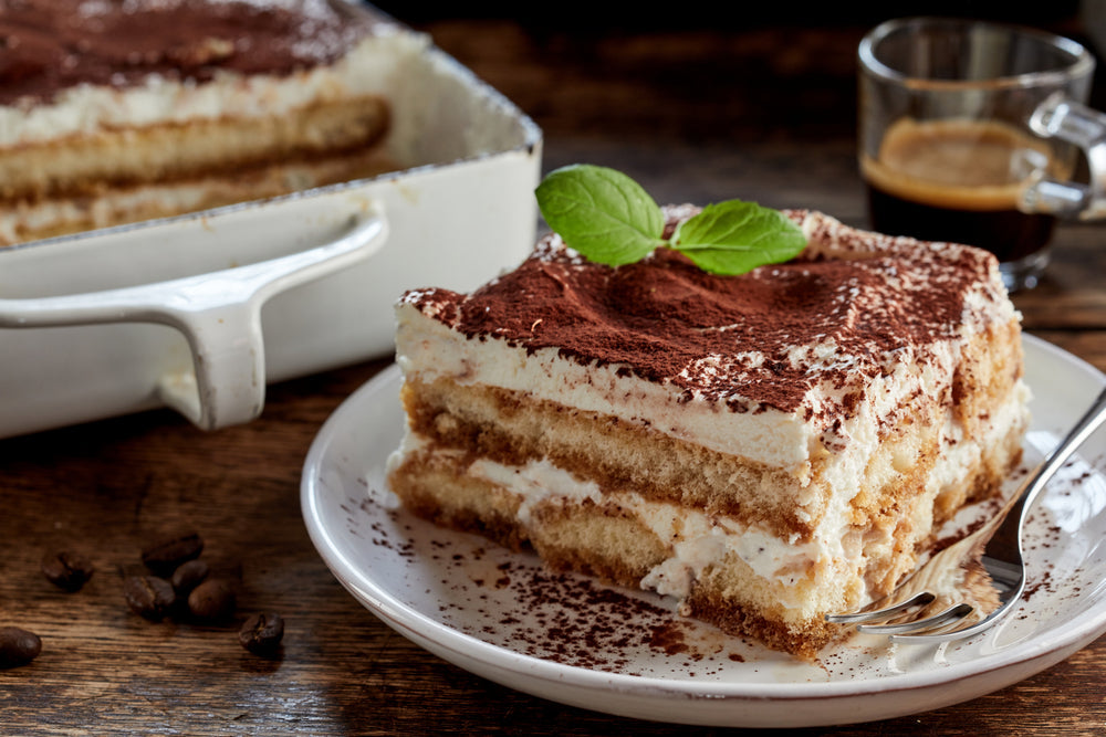 A slice of tiramisu topped with cocoa powder and a mint leaf sits on a white plate next to a serving dish, with coffee beans scattered nearby and a cup of espresso in the background.