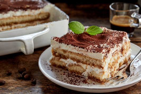 A slice of tiramisu topped with cocoa powder and a mint leaf sits on a white plate next to a serving dish, with coffee beans scattered nearby and a cup of espresso in the background.