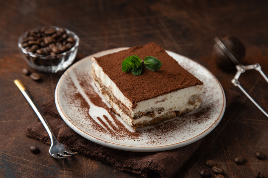 A slice of tiramisu topped with cocoa powder and a mint leaf sits on a white plate, accompanied by a bowl of coffee beans and a fork on a brown napkin, against a rustic wooden background.