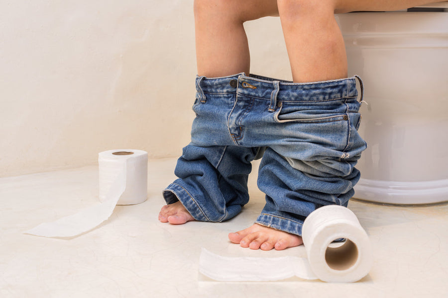 A child sits on a toilet with their jeans around their ankles, surrounded by two rolls of toilet paper on a light-colored bathroom floor.