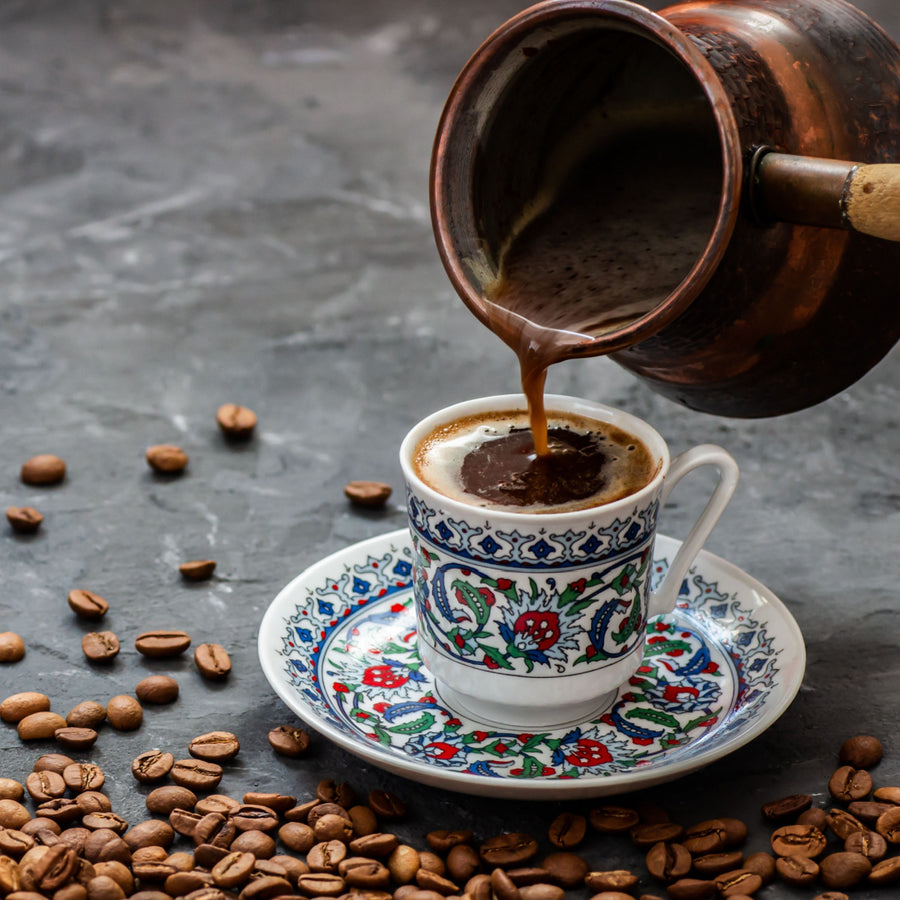A copper pot pours rich coffee into a decorative cup on a patterned saucer, surrounded by scattered coffee beans on a dark surface.