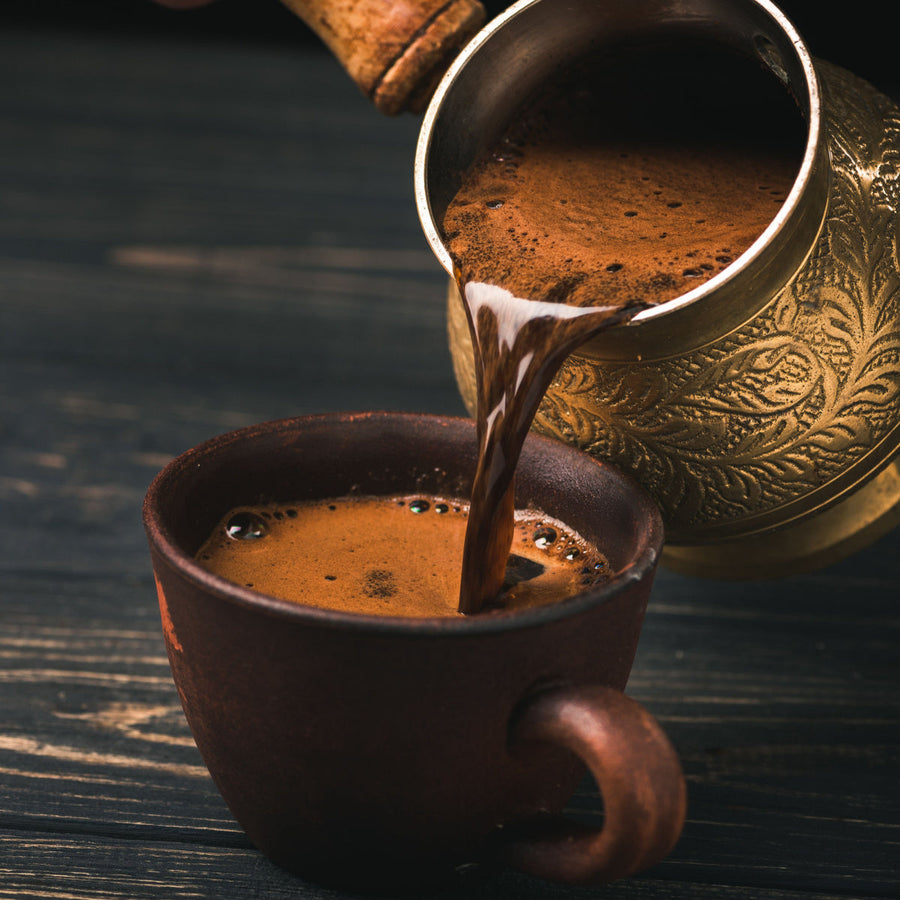 A brass pot pours rich, dark coffee into a rustic brown cup, with bubbles forming on the surface, set against a dark wooden background.