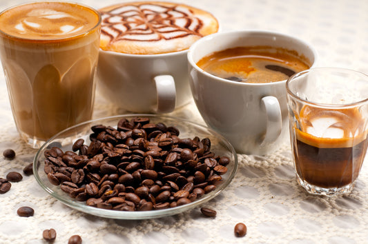 Three cups of coffee, including a latte and a cappuccino with decorative foam, sit on a textured surface alongside a plate of coffee beans and a small glass of espresso.