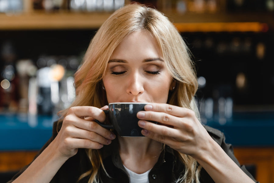 A woman with long blonde hair closes her eyes and enjoys a warm drink from a black cup while sitting in a cozy café setting.