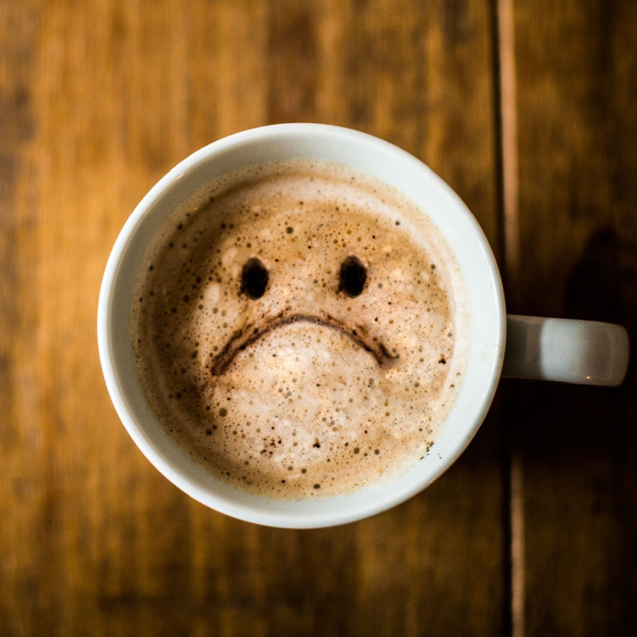 A white coffee cup sits on a wooden table, with frothy coffee displaying a sad face made of milk and coffee, featuring a frown and two dark spots for eyes.