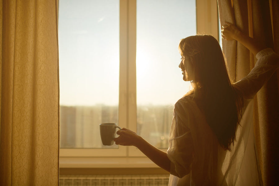 A woman in a white shirt stands by a window, holding a cup of coffee and pulling back the curtain to let in the morning sunlight, with a cityscape visible in the background.