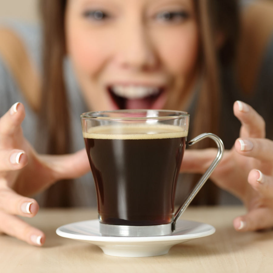 A woman with a joyful expression reaches eagerly for a glass of coffee on a white saucer, with a blurred background suggesting a cozy indoor setting.