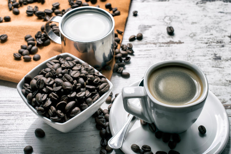 A cup of coffee sits on a white plate next to a bowl of coffee beans, with a silver milk container and scattered beans on a wooden surface, creating a cozy coffee setting.