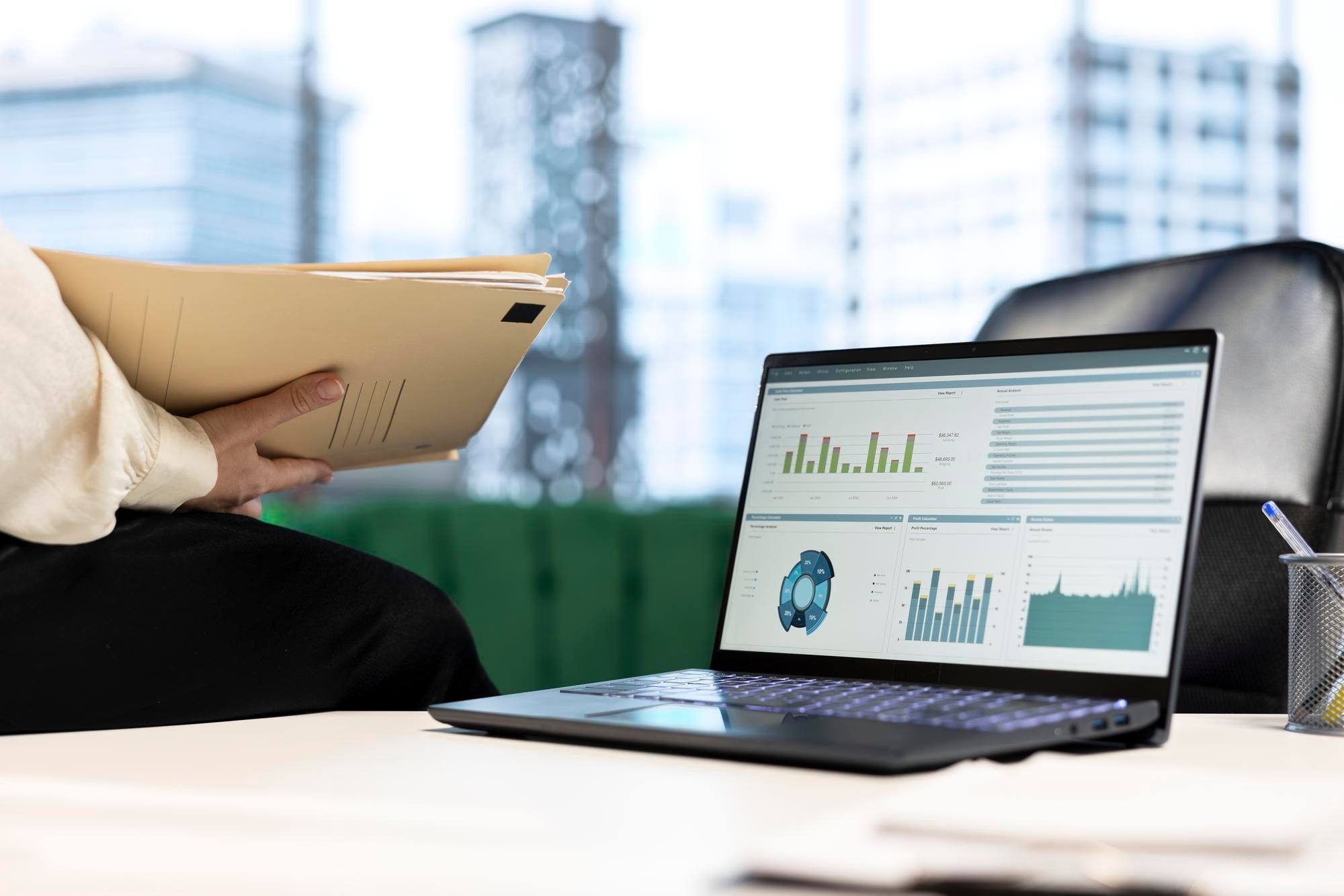A person holding a folder sits next to a laptop displaying various data charts and graphs, with a modern office background featuring large windows and city buildings.