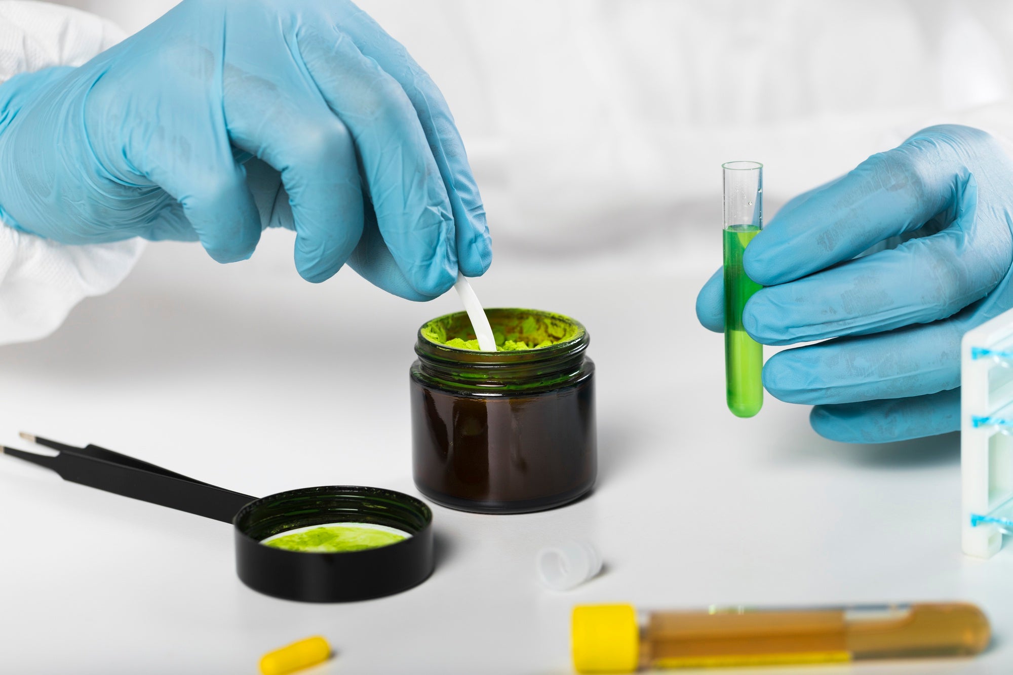 A gloved hand is using a spatula to mix a green powder in a dark jar, while another hand holds a test tube containing bright green liquid, with various lab tools visible on a white surface.