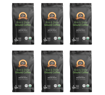 Six black bags of Alex's Low-Acid Organic Coffee™ - Rise and Shine Fresh Ground (12oz) by Alex's Low Acid Coffee, with orange/brown circular logos, marked as gentle on stomach, low acid, medium roast, USDA organic, and fair trade.