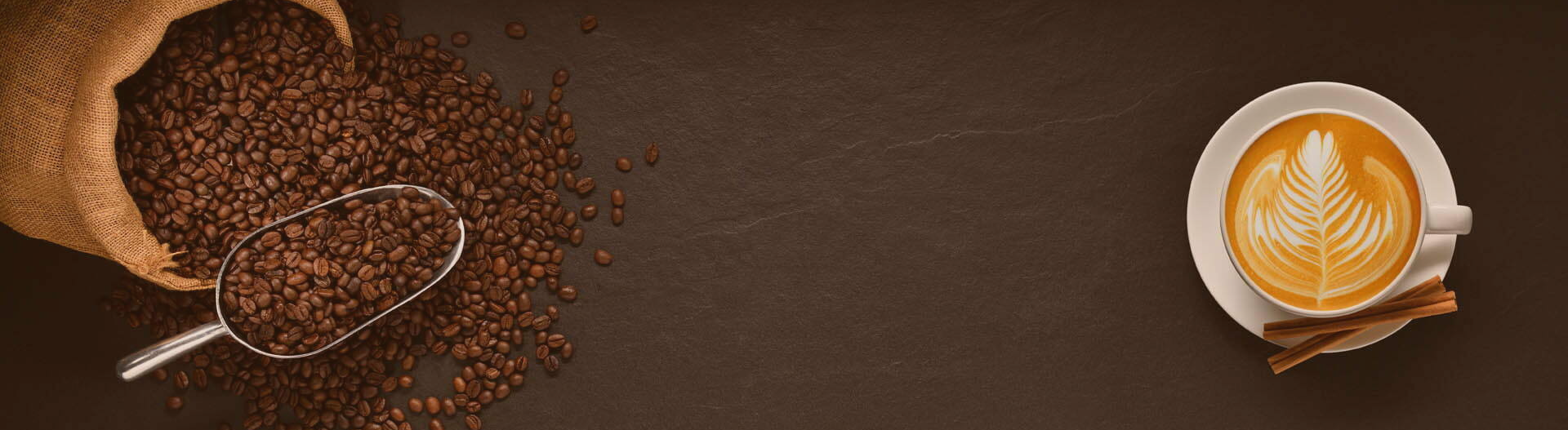 A burlap sack spills coffee beans onto a dark surface, next to a metal scoop. On the right, a cup of cappuccino with latte art and two cinnamon sticks rests on a saucer.